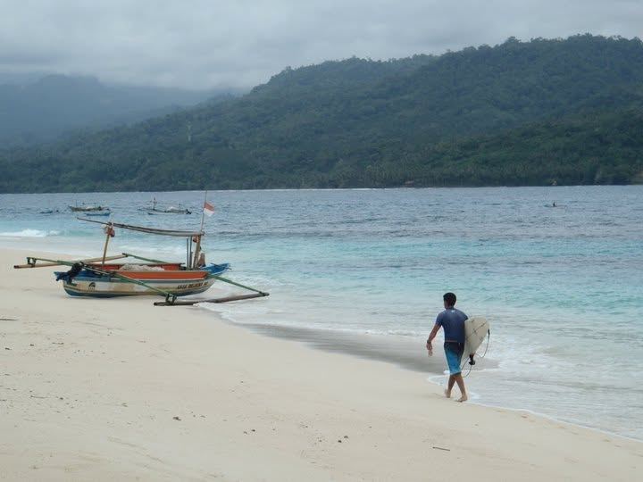 Kevin walking up the beach with his board, past an outrigger boat